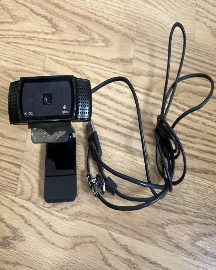 Logitech HD 1080p USB webcam placed on a wooden floor, commonly used as podcast equipment for video recordings or livestreams.