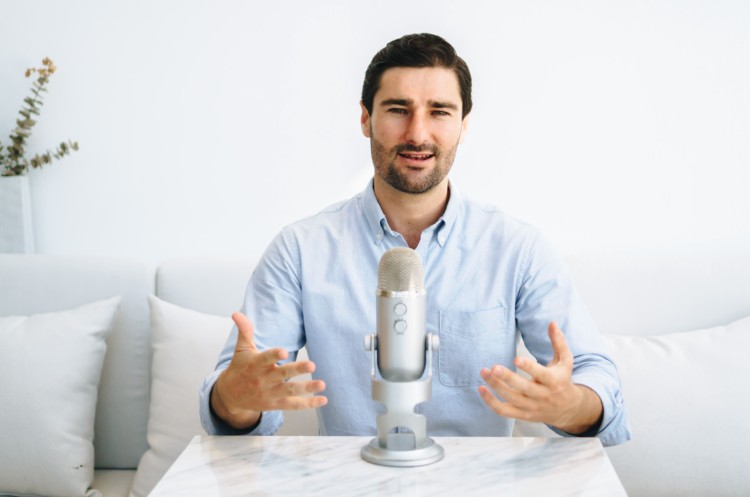 Man sitting at a table with a microphone, gesturing as he speaks during a podcast host recording session.