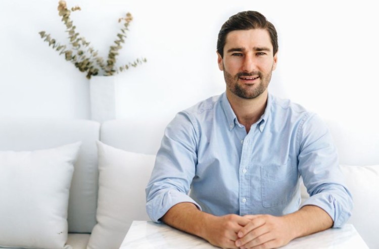 Liam Austin sitting at a table in a light blue button-down shirt, demonstrating the likely attire for male guest speaker appearances.
