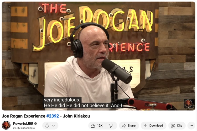 Joe Rogan speaking into a microphone in a studio with a neon logo sign and wood-paneled walls, showing a bold podcast space setup.