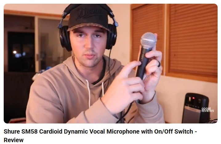 Reviewer holding and pointing to a Shure SM58 vocal microphone while wearing headphones, demonstrating features during an in-depth review.